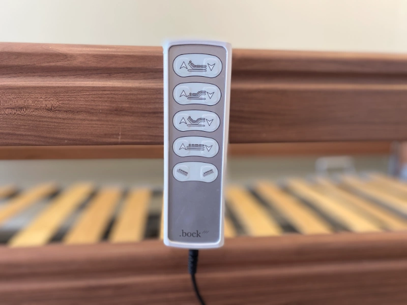 Elektromos betegágy távirányító közelről, háttérben lamellás fekvőfelület